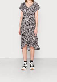 Kvinna i en svart blommig midikjol med korta ärmar, som matchar med svartvita högsnörade sneakers, står mot en enfärgad bakgrund.
