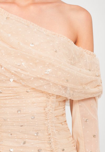 Vrouw in een sheer, off-shoulder beige jurk met kleine glanzende pailletten en gerimpelde stofdetails.