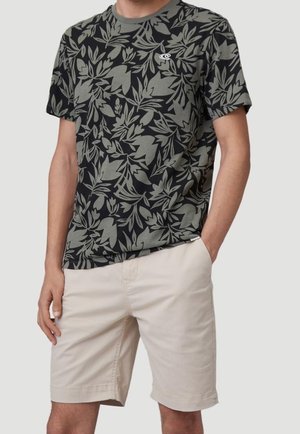 Uomo che indossa una t-shirt nera e verde oliva con motivo floreale e pantaloncini beige al ginocchio, una mano in tasca, su sfondo neutro.