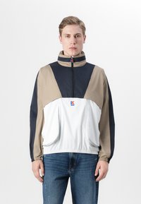 Lett anorakkjakke i beige, marineblå og hvit, med høy krage, halv glidelås og elastiske mansjetter. Logo på brystet.