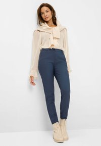 Pantalons ajustés bleu marine avec une fermeture éclair à l'avant et une texture lisse, associés à un chemisier beige transparent avec des détails en volants et des manches évasées.