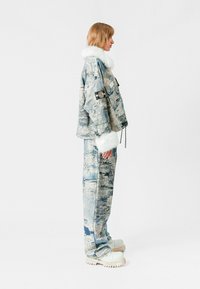 Chaqueta de mezclilla azul y blanca con un cuello de piel, combinada con pantalones a juego y zapatos de plataforma gruesos en blanco roto. Vista lateral.