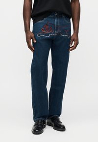 Σκούρα μπλε τζιν από denim με μοναδικό σχέδιο και κόκκινες και ασημένιες διακοσμήσεις στις πίσω τσέπες. Κανονική εφαρμογή με ίσια γραμμή και κλασικό στυλ.