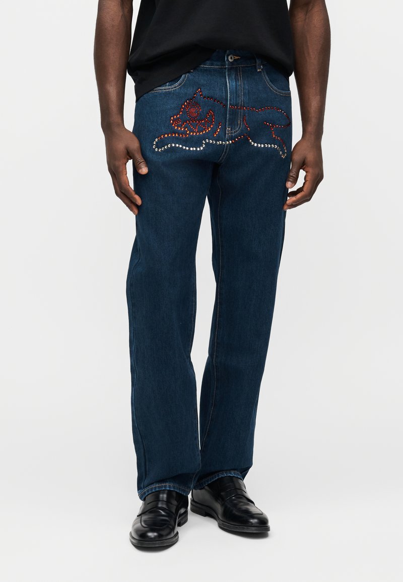 Σκούρα μπλε τζιν από denim με μοναδικό σχέδιο και κόκκινες και ασημένιες διακοσμήσεις σ τις πίσω τσέπες. Κανονική εφαρμογή με ίσια γραμμή και κλασικό στυλ.