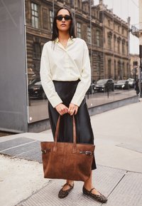 Borsa a tracolla marrone con due manici, realizzata in materiale testurizzato. Il modello indossa una camicia gialla chiaro con bottoni e una gonna nera in satin, con ballerine a stampa animale.