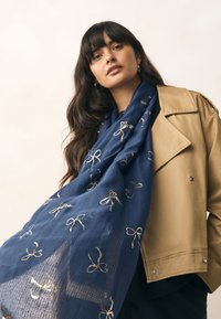 Sciarpa blu navy in tessuto leggero, con motivi di fiocchi dorati. Indossata con una giacca color cammello, presenta una texture morbida e un design drappeggiabile.