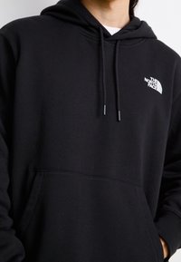 Personne portant un sweat à capuche noir avec poches avant et le logo blanc The North Face sur la poitrine, visage et mains partiellement visibles.