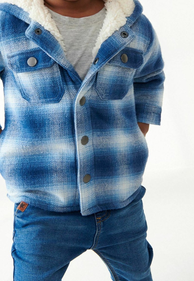 Enfant portant une veste à carreaux bleue et blanche avec doublure en polaire et un jean bleu, les mains glissées dans les poches de la veste.