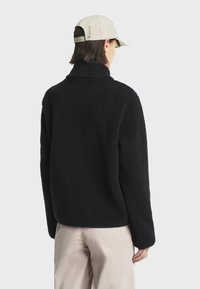 Μαύρο φλις μπουφάν με ψηλό γιακά, που επιδεικνύει μια απαλή υφή. Φοριέται με μια μπεζ καπελάκι, που διαθέτει ένα διακριτικό λογότυπο στο πλάι.