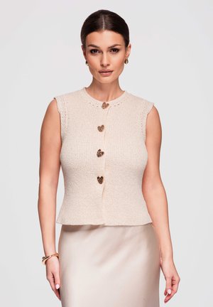 Vrouw met donker haar, goudkleurige oorringen, een mouwloze beige gebreide top met hartvormige knopen en een lichtbeige rok.