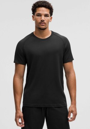 Mand med kort krøllet hår iført en sort, kortærmet T-shirt med rund hals og sorte bukser mod en ensfarvet grå baggrund.