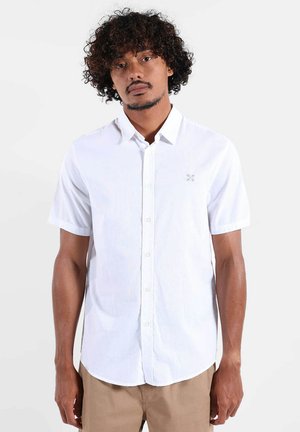 Homme aux cheveux bouclés portant une chemise blanche à manches courtes et un pantalon beige, debout devant un fond clair uni.