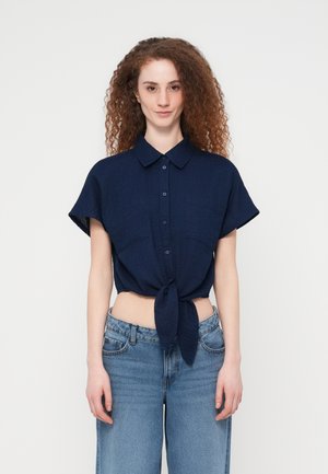 Femme aux cheveux roux bouclés portant une chemise bleu marine à manches courtes boutonnée, nouée à la taille, et un jean bleu taille haute, debout devant un fond blanc.