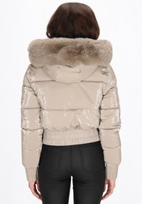 Jachetă matlasată bej cu finisaj lucios, guler din blană artificială, tiv și manșete elastice, și design cropped, văzută din spate.