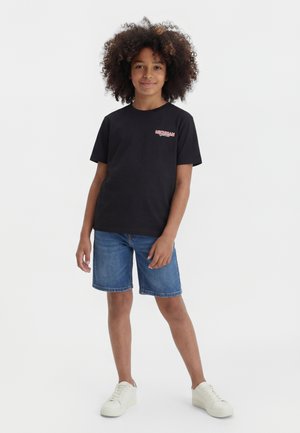 Kind mit lockigem Haar, das ein schwarzes T-Shirt mit dem Schriftzug Michigan, Jeansshorts und weiße Turnschuhe trägt, steht mit einer entspannten Hand.