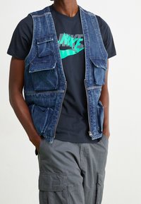 Osoba v tmavém tričku Nike, rozepnuté denimové vesta s mnoha kapsami a šedých cargo kalhotách se zapíchnutýma rukama v kapsách.