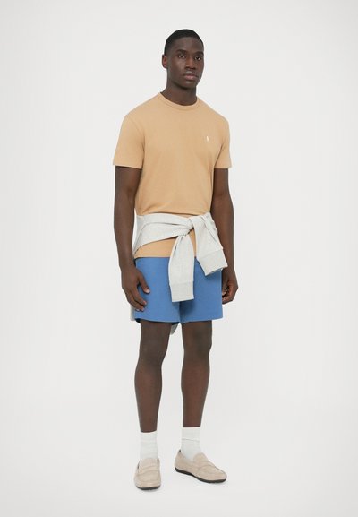 Pamut bézs póló logóval, szürke pulóver deréknál megkötve, kék rövidnadrág és bézs csúszásmentes cipő fehér zoknival.