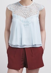 Vaaleansininen yläosa, jossa on pitsiyksityiskohtia kukkakuviolla, yhdistettynä burgundinpunaisiin korkeavyötäröisiin shortseihin, joissa on edessä poimutuksia.