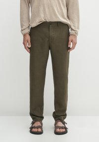 Pantaloni in lino verde oliva con vestibilità dritta, tasche frontali e chiusura con un bottone, abbinati a sandali marroni.