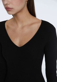 Schwarzes, geripptes Langarmoberteil mit V-Ausschnitt. Das Material wirkt glatt mit einem strukturierten Muster, das die taillierte Silhouette betont.