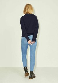 Merino villatakki, vaaleansiniset kapeat farkut, mustat nilkkurit ruskeilla pohjilla. Malli katsoo pois päin, kädet ristissä takana. Vaalea tausta.