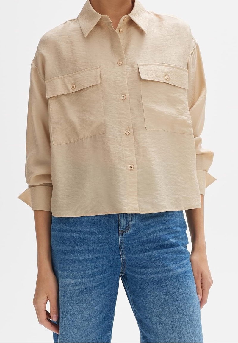 Chemise beige boutonnée avec deux poches poitrine et manches longues, assortie à un jean bleu sur une personne debout devant un fond uni.