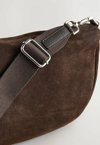 Dunkelbraune Wildleder-Handtasche mit strukturierter Oberfläche, verstellbarem Stoffriemen und silbernen Beschlägen für den Verschluss und die Riemenverbindung.