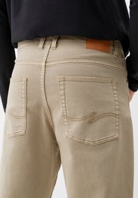 Pantalones de mezclilla beige con una textura suave, que presentan dos bolsillos traseros, detalles de costura sutiles y una etiqueta de marca de cuero sobre la cinturilla.