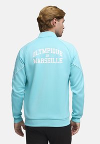 Chaqueta deportiva azul claro con texto blanco "OLYMPIQUE DE MARSEILLE" en la espalda, puños acanalados y tejido suave. Presenta un cuello con cremallera y paneles laterales.