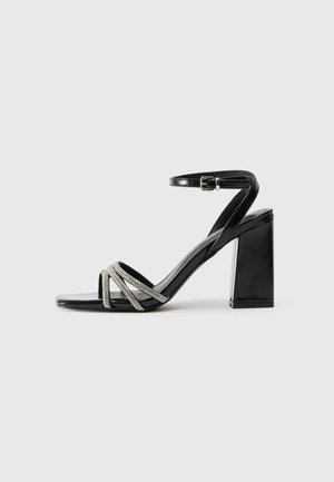 Sandale noire à talon haut avec un talon bloc, dotée d'une bride avec des accents métalliques et de trois bandes croisées au niveau des orteils.