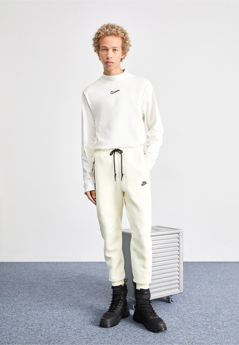 Junge Person mit lockigem Haar, die einen weißen Nike-Sweatshirt und Jogginghosen mit schwarzen Stiefeln trägt, steht neben einem kleinen silbernen Rollkoffer auf grauem Boden.