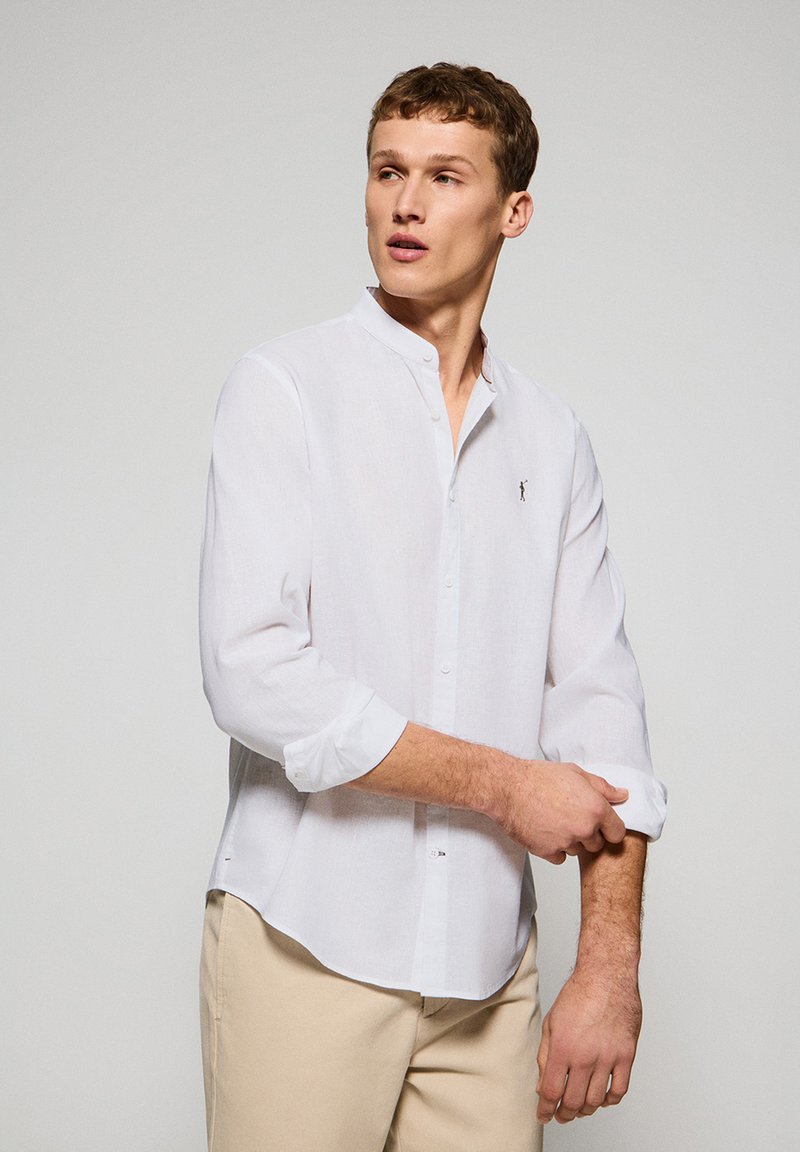 Vit skjorta med mandarin-krage, långa ärmar rullade vid ärmsluten och en liten broderad logotyp på bröstet. Lättviktsmaterial.