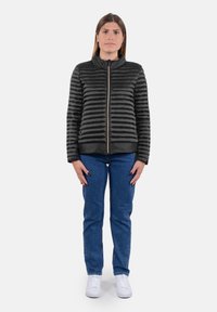 Giacca nera trapuntata con colletto alto, zip completa e righe orizzontali. Abbinata a jeans di denim blu e scarpe da ginnastica bianche.