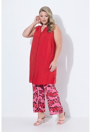 Nainen pukeutunut hihattomaan punaisen napitettuun mekkoon leveälahkeisten kukkakuosisten vaaleanpunaisten ja punaisten housujen päällä, vaaleanpunaiset korolliset sandaalit jalassa, poseeraa valkoisen seinän edessä.