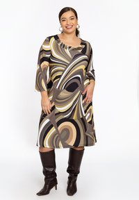 Langärmliges Kleid mit abstrakten Wirbeln in Braun, Creme und Gold, mit einem mit Perlen verzierten Ausschnitt, kombiniert mit kniehohen schwarzen Stiefeln.