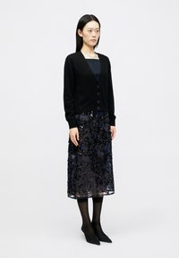 Cardigã preto com decote em V e fecho de botões, usado sobre uma blusa azul-marinho e uma saia preta semi-transparente, com contas e lantejoulas, combinada com meias pretas e sapatos.