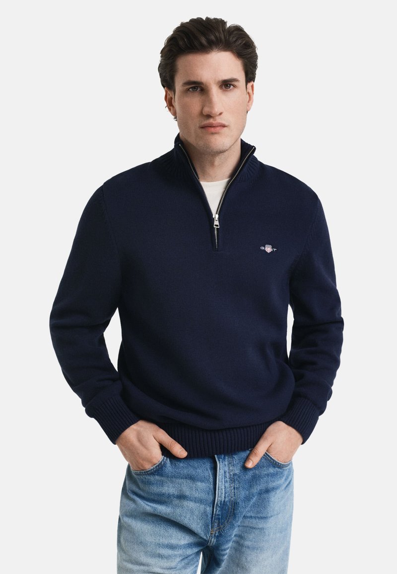 Dunkelblauer Strickpullover mit einem Reißverschlusskragen, gerippten Bündchen und Saum, kombiniert mit hellblauen Jeans. Verfügt über kleine Logodetails auf der Brust.