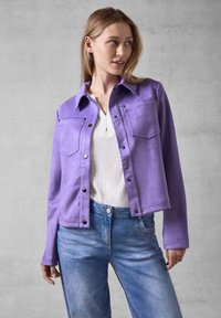 Veste en daim violet avec poches avant et fermeture à boutons-pression, accompagnée d'un haut blanc et d'un jean bleu, sur fond neutre.