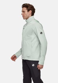 Helle mintgrüne Softshelljacke mit hohem Kragen, seitlichen Taschen und schwarzem Logo am Ärmel, mit elegantem, taillierten Design.