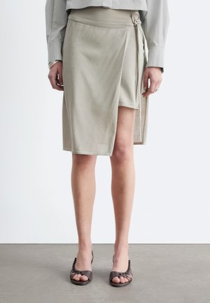 Osoba oblečená v béžovej zavinovacej sukni s bočnou stuhou, sivom tričku s dlhým rukávom a čiernych otvorených podpätkoch stojaca na šedom podlahe.