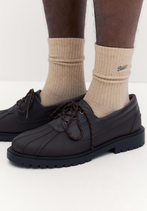 Sötétbarna bőr fűzős cipők bézs bordázott zoknival, sötét bőrű lábakon, egyszínű fehér háttér előtt.