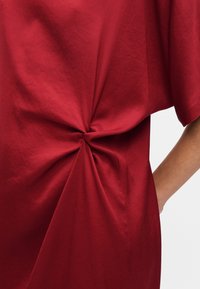 Haut en coton rouge avec un détail noué torsadé à la taille, présentant une coupe ample et des épaules tombantes. Texture lisse et design minimaliste.