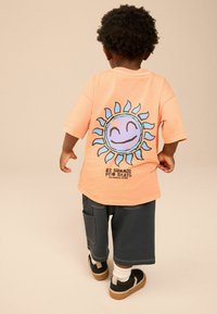 Lichtoranje t-shirt met een blauwe en zwarte zonafbeelding op de achterkant, gecombineerd met donkere shorts en zwarte sneakers met witte sokken.