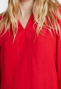 Persona bionda che indossa una maglietta rossa brillante a V, con capelli lunghi e leggermente ondulati che cadono sulle spalle.