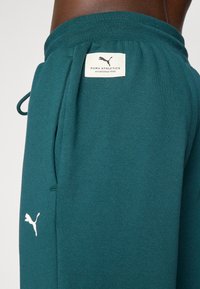 Teal sweatpants lavet af blødt stof, med snøre i taljen, sidelommer og et hvidt logopatch med teksten "PUMA ATHLETICS."