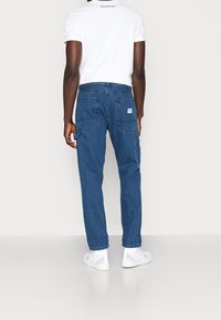 Vaqueros de mezclilla azul oscuro de corte recto, con dos bolsillos traseros y una etiqueta con el logo en blanco. Combinados con una camiseta blanca lisa.