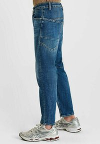 Zijaanzicht van een persoon die blauwe denim jeans en zilveren sport sneakers draagt, staande op een witte achtergrond.