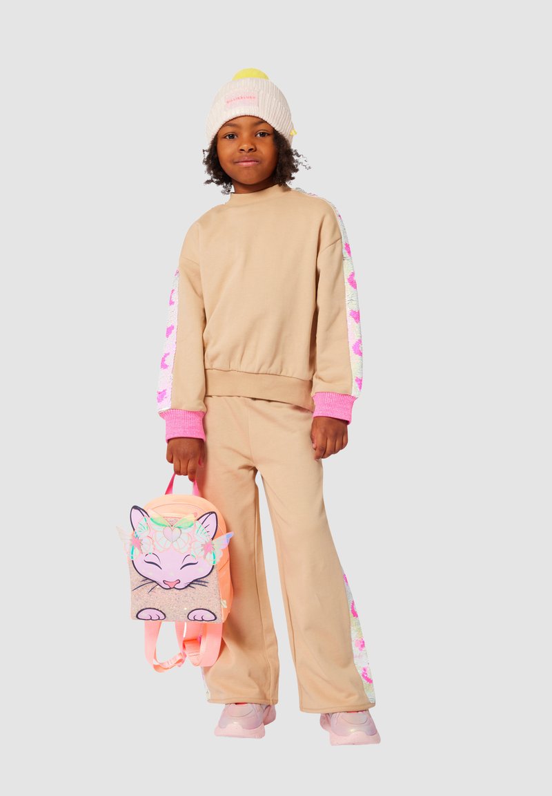 Een jong kind poseert in een beige trainingspak met roze accenten en een lichtgekleurde beanie. Ze houden een roze rugzak met katten-thema vast.