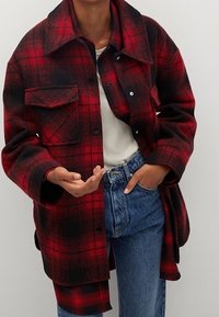 Rød og sort ternet oversized flannel skjortejakke med krave, frontlomme og trykknapper, lagt over en hvid top og blå jeans.