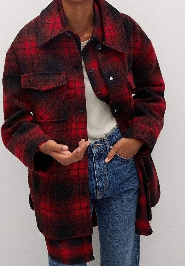 Rød og sort ternet oversized flannel skjortejakke med krave, frontlomme og trykknapper, lagt over en hvid top og blå jeans.